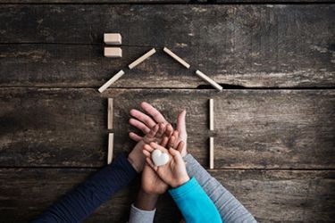 Photographie de mains prenant un objet en forme de cœur entouré de kapla représentant une maison